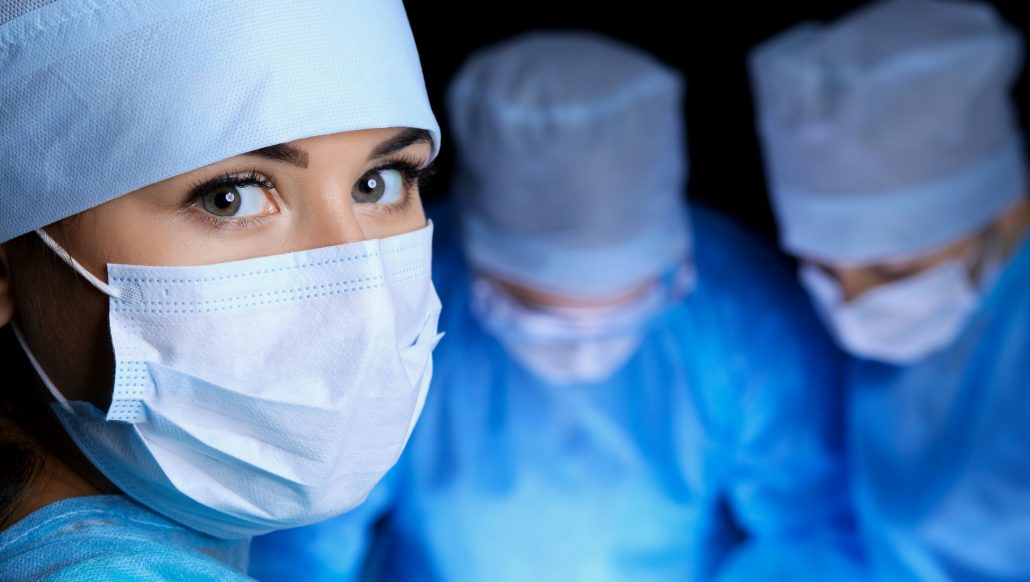 Undersköterska tittar in i kameran medans två personer opererar i bakgrunden