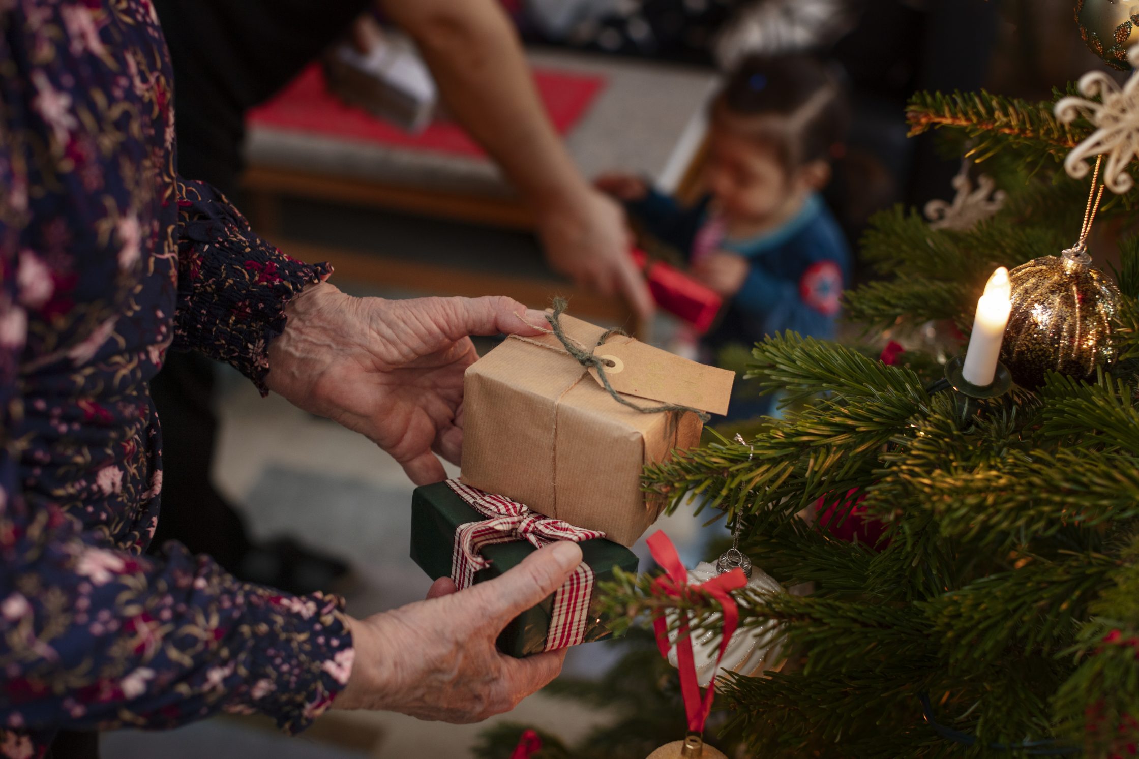 En äldre hand som håller i en julklapp intill julgranen.