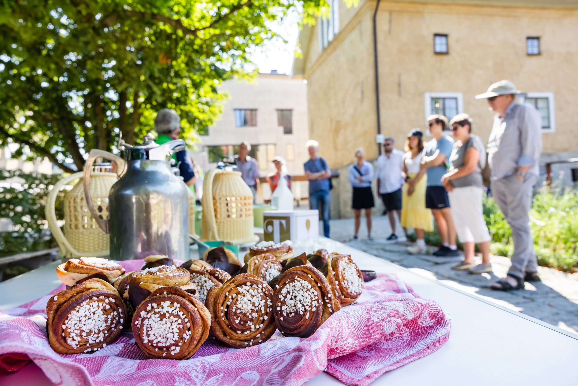 Ett fat med kakor och bullar. I bakgrunden syns en grupp människor på fikavandring, framför Alingsås museum.