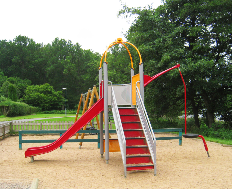 Lekplatsmodul med rutschkana och en trappa. Färgerna röd och gul dominerar. Gungställning och träd i bakgrunden.